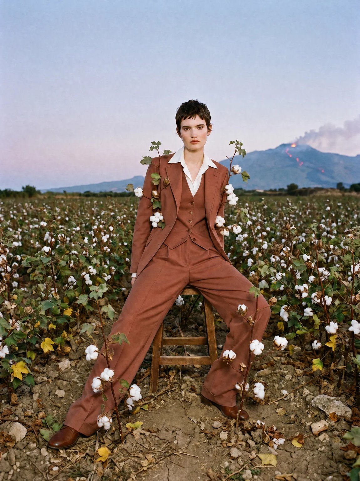 Man in a terracotta-colored suit holding cotton branches, sitting in a cotton field at the foot of Mount Etna